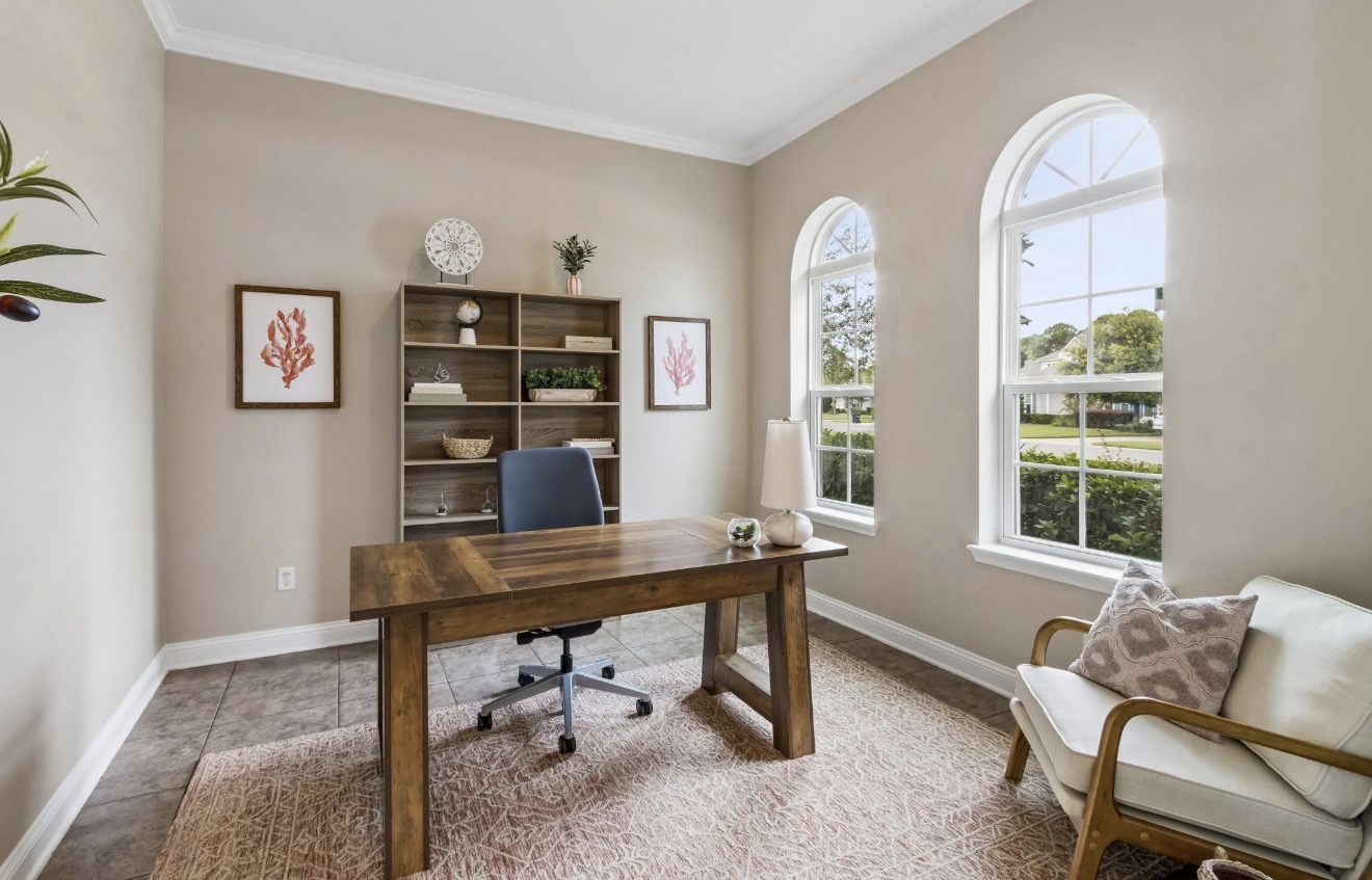Home office staged with a wood desk, navy chair, coral art prints, and arched windows
