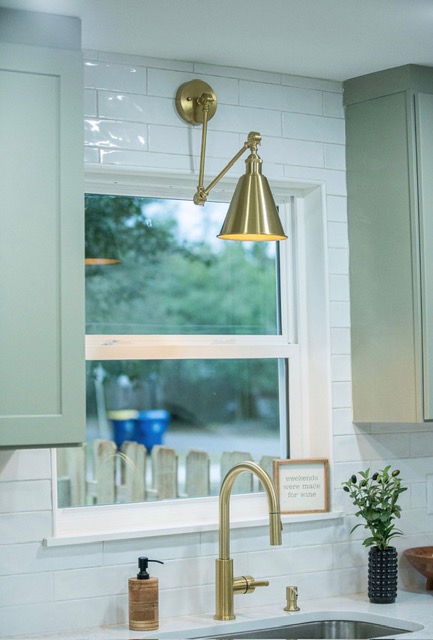 Sage green cabinetry with brass sconce, subway tile, and brass bar faucet