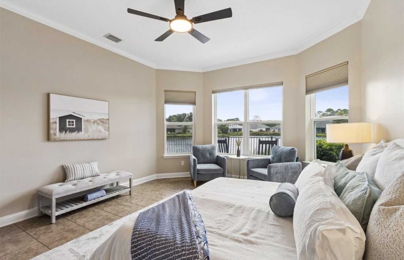 Primary bedroom staged with neutral linens, blue accent chairs, and water views